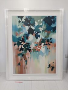 An original painting on canvas of a leafy canopy scene in soft colours of green, blue and peachy ochre, in a bespoke off white frame. set against light coloured wall.
