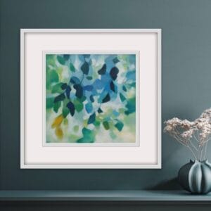 An original painting on canvas of tree leaves in spring like shades of blue and green, with a hint of yellow, in a bespoke off white frame set against a deep grey green grey wall with a shelf below.