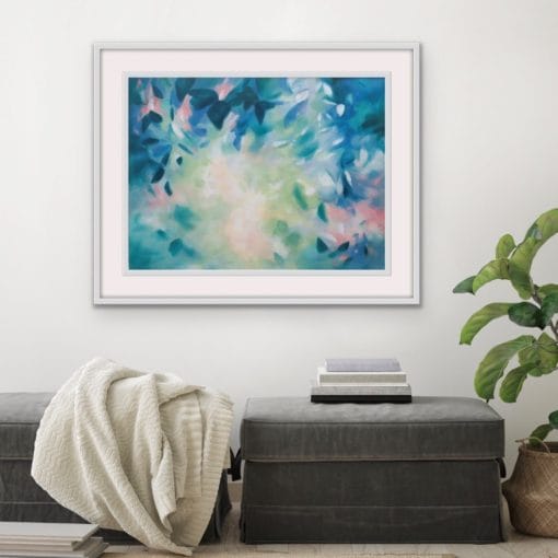 An original painting on canvas in Soft Green and Blue colours with pops of pink, of a leafy tree canopy with a Spring like feel in a bespoke with frame. Set against a white wall over a grey velvet footstool with plant and throw.